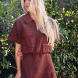 Woman wearing a brown jumpsuit standing in front of green foliage