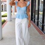 Woman wearing a blue checkered top and white pants standing on a sidewalk.
