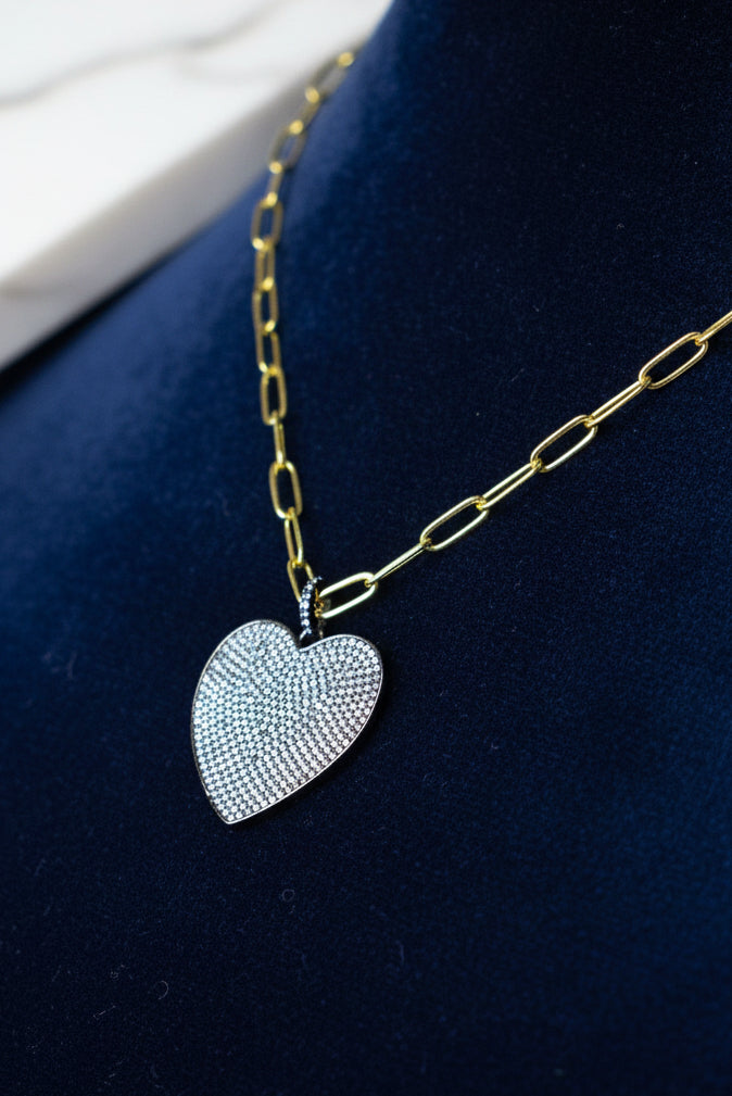 A gold-colored chain necklace with a black and silver oversized heart-shaped pendant on a mannequin bust.