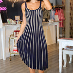 A woman stands in a boutique wearing a sleeveless navy Irecia Sweater midi dress with vertical white stripes and white heeled sandals.