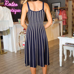 A woman stands in a boutique wearing a sleeveless navy Irecia Sweater midi dress with vertical white stripes and white heeled sandals.