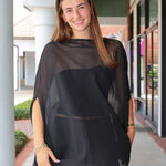A woman wearing a sheer black mesh poncho-style top with a solid black underlayer, standing in a covered outdoor walkway.