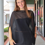 A woman wearing a sheer black mesh poncho-style top with a solid black underlayer, standing in a covered outdoor walkway.