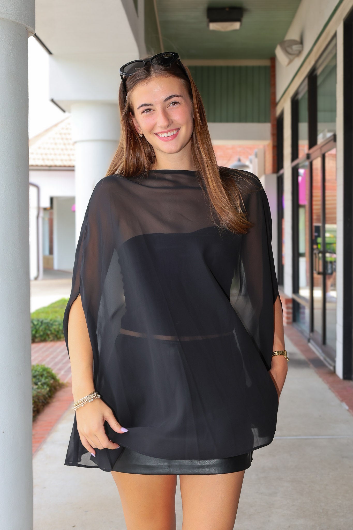 A woman wearing a sheer black mesh poncho-style top with a solid black underlayer, standing in a covered outdoor walkway.