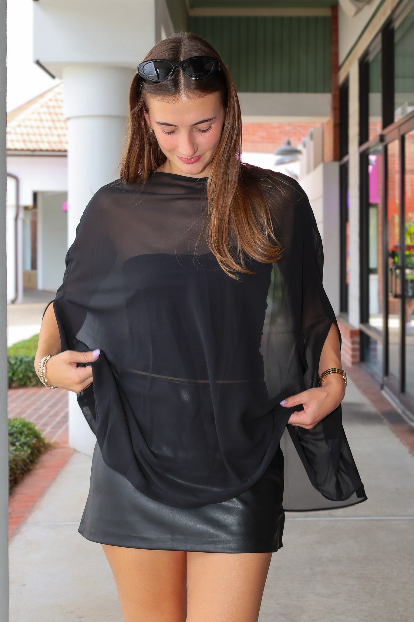 A woman wearing a sheer black mesh poncho-style top over a black faux-leather mini skirt, standing on an outdoor walkway.