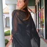 A woman wearing a sheer black long-sleeve open-front cover-up or poncho, photographed from the back in an outdoor walkway setting.