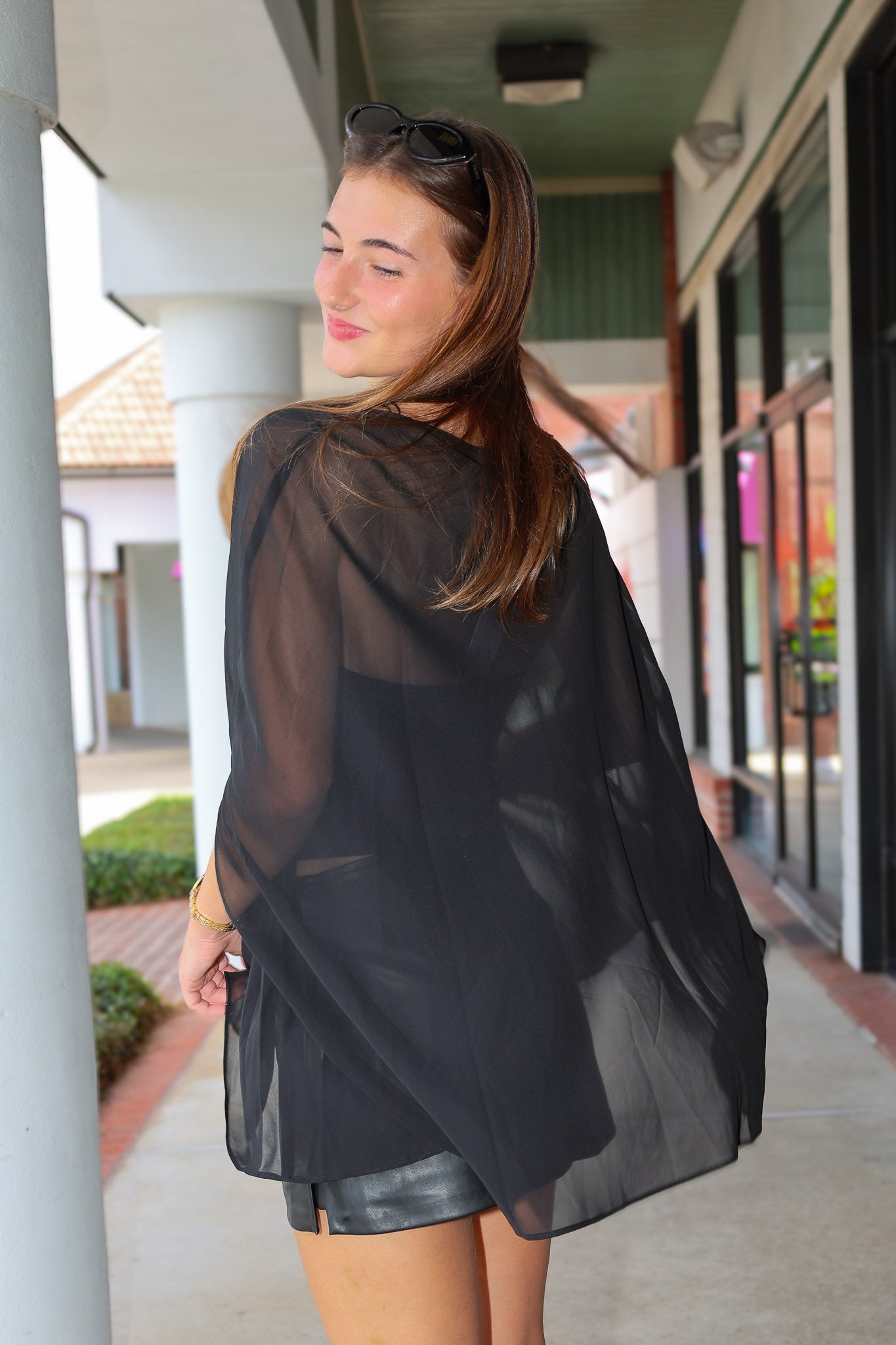 A woman wearing a sheer black long-sleeve open-front cover-up or poncho, photographed from the back in an outdoor walkway setting.