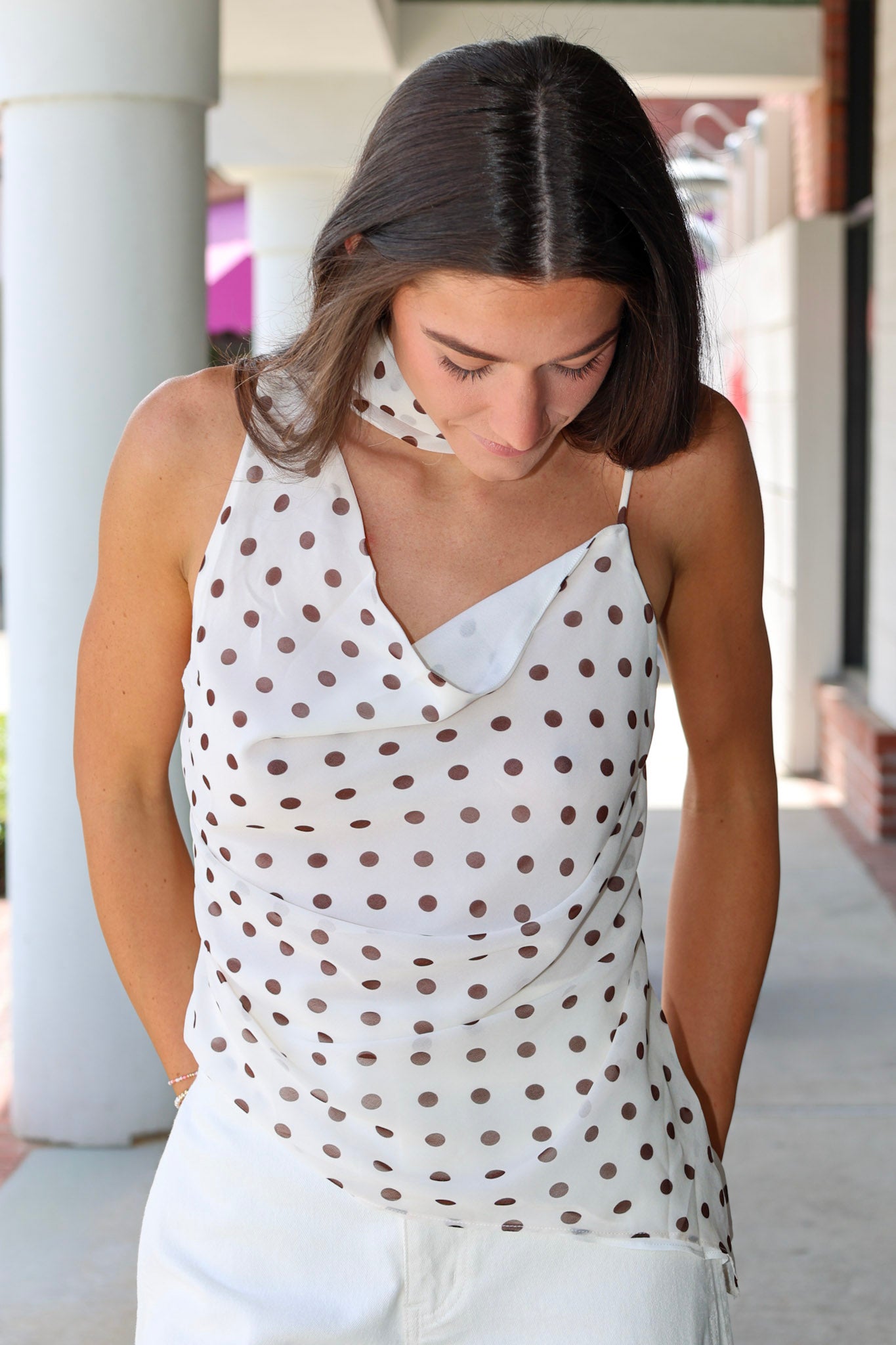 Woman wearing a white sleeveless top with brown polka dots and white pants outdoors.