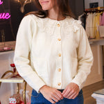 A woman wearing a cream-colored knit cardigan with a large lace Peter Pan collar and  buttons, paired with a denim mini skirt, standing in Rodeo boutique.