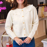 A woman modeling a cream-colored knit cardigan with a large lace Peter Pan collar and gold buttons, paired with a blue denim mini skirt in a boutique setting.