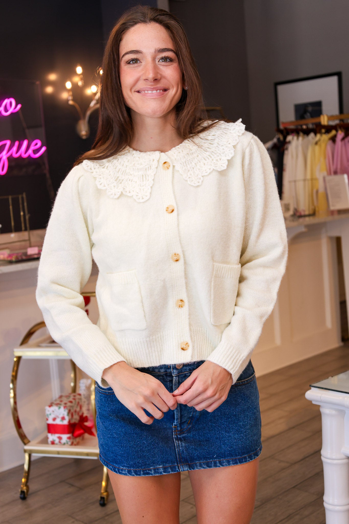 A woman modeling a cream-colored knit cardigan with a large lace Peter Pan collar and gold buttons, paired with a blue denim mini skirt in a boutique setting.