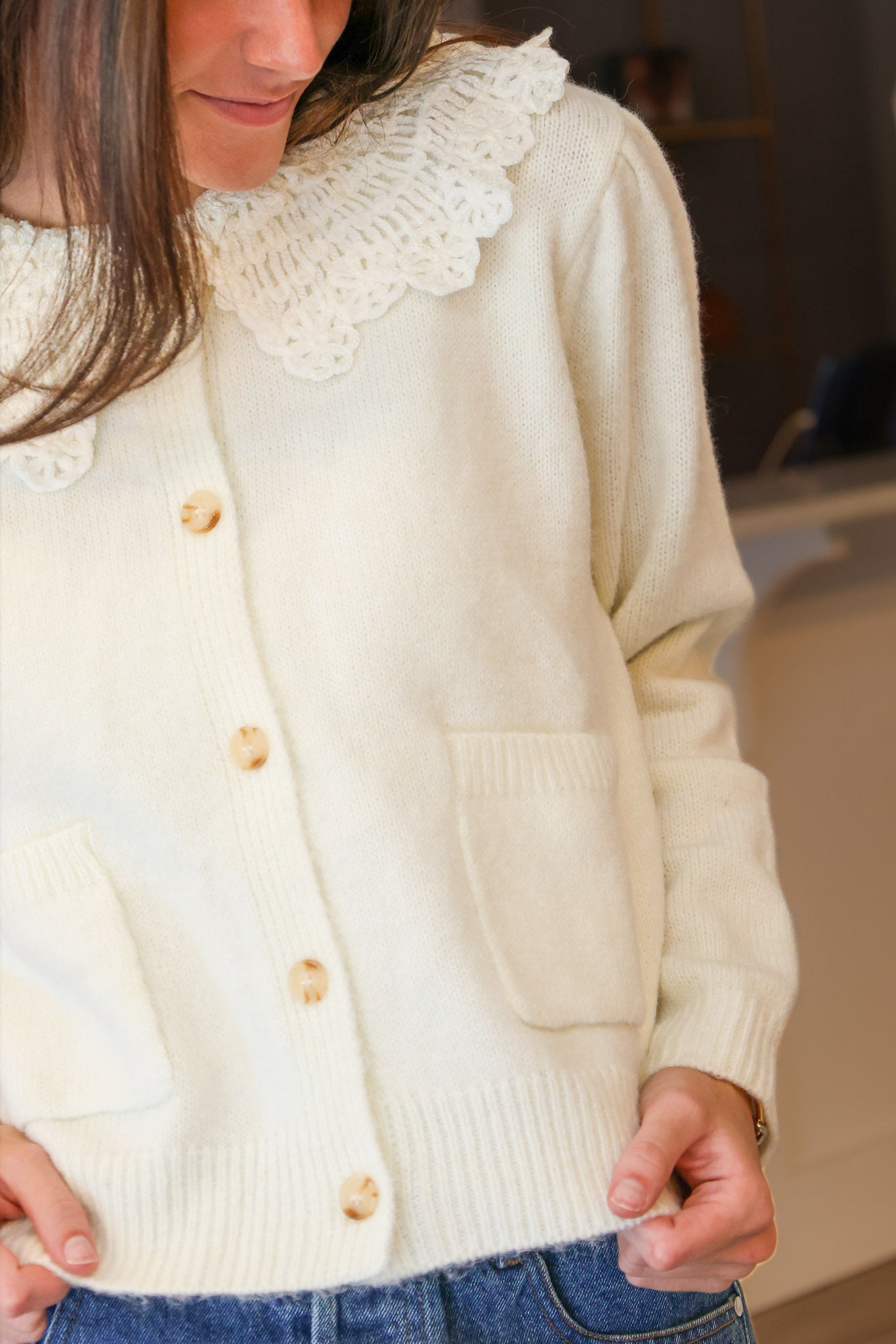 A close-up of a person wearing a cream-colored knit cardigan with a large white lace Peter Pan collar and light wood buttons, paired with a denim skirt at Rodeo Boutique..
