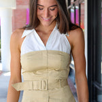 A woman wearing a beige and white halter top with a structured bust, front pleats, and a matching belt, paired with blue jeans in an outdoor walkway setting.