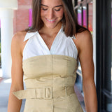 A woman wearing a beige and white halter top with a structured bust, front pleats, and a matching belt, paired with blue jeans in an outdoor walkway setting.