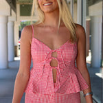 A smiling blonde woman wearing a red and white gingham two-piece set with a tie-front peplum top and matching fitted pants, standing in a covered walkway.