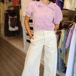 A woman wearing a light purple lavender short-sleeve ribbed knit cardigan with a ruffled collar and button-front closure, paired with white pants and sunglasses, standing in Rodeo Boutique.