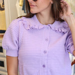 A woman wearing a light purple short-sleeve ribbed knit cardigan with a ruffled collar and button-front closure, paired with white pants and sunglasses, standing in Rodeo Boutique.