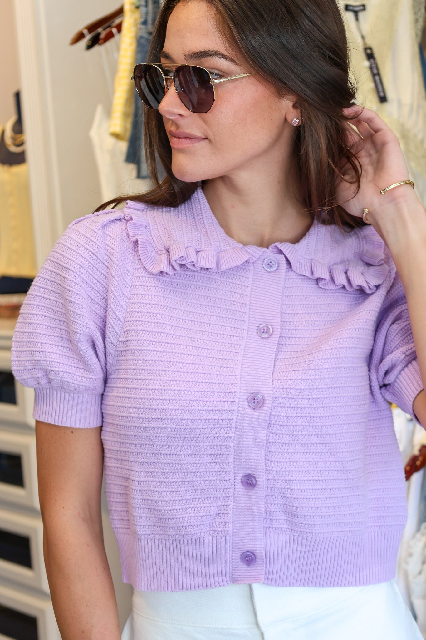 A woman wearing a light purple short-sleeve ribbed knit cardigan with a ruffled collar and button-front closure, paired with white pants and sunglasses, standing in Rodeo Boutique.