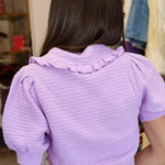 A woman wearing a light purple short-sleeve ribbed knit cardigan with a ruffled collar and button-front closure, paired with white pants and sunglasses, standing in Rodeo Boutique.