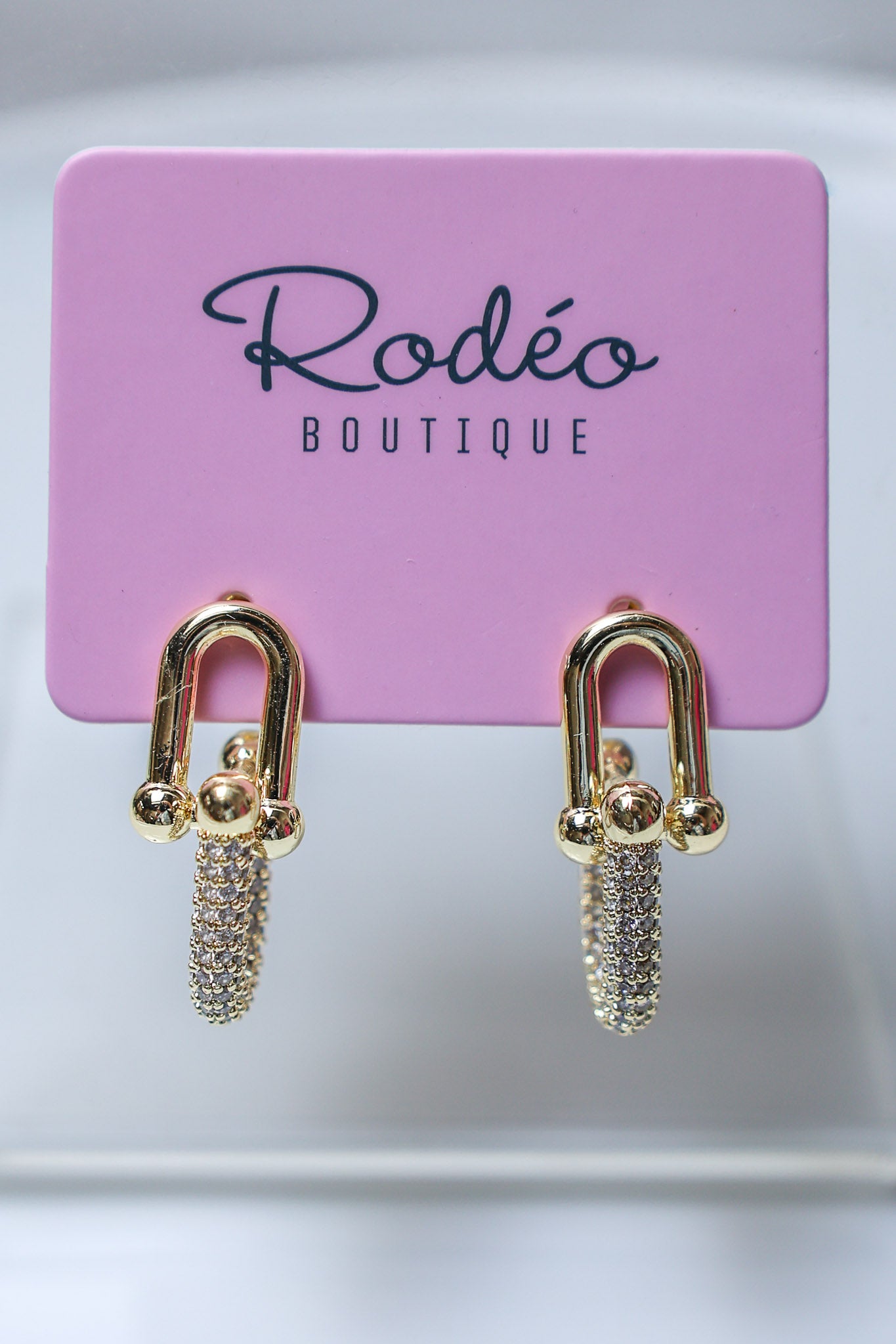 A pair of gold-tone drop earrings with pavé crystal chains, displayed on a pink card labeled 'Rodéo BOUTIQUE'.