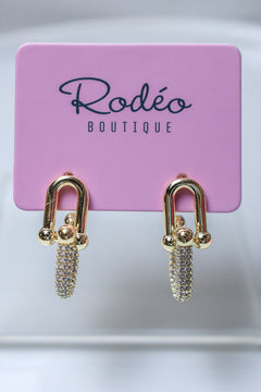 A pair of gold-tone drop earrings with pavé crystal chains, displayed on a pink card labeled 'Rodéo BOUTIQUE'.