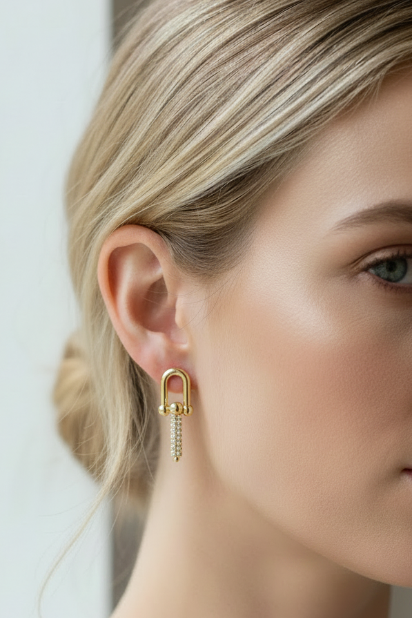 A close-up of a woman's ear wearing a gold hoop earring with a dangling chain of small clear stones, shown against her blonde hair and face.