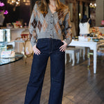 Woman in a patterned argyle cardigan and dark jeans standing in an indoor setting with tables and chairs.