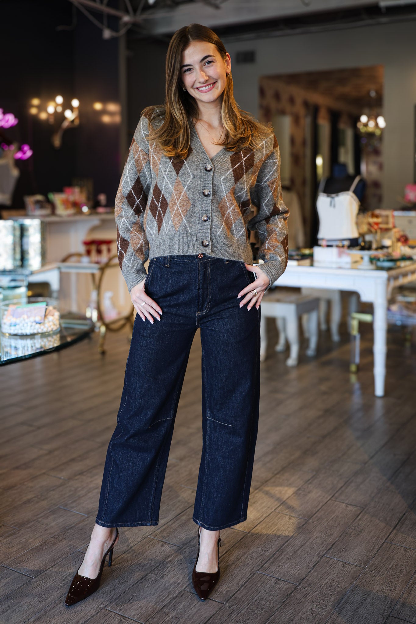 Woman in a patterned argyle cardigan and dark jeans standing in an indoor setting with tables and chairs.