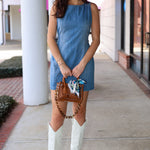 Woman in a blue denim dress with white boots standing on a sidewalk.