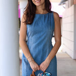 Woman wearing a sleeveless denim dress holding a brown clear handbag in an outdoor setting.