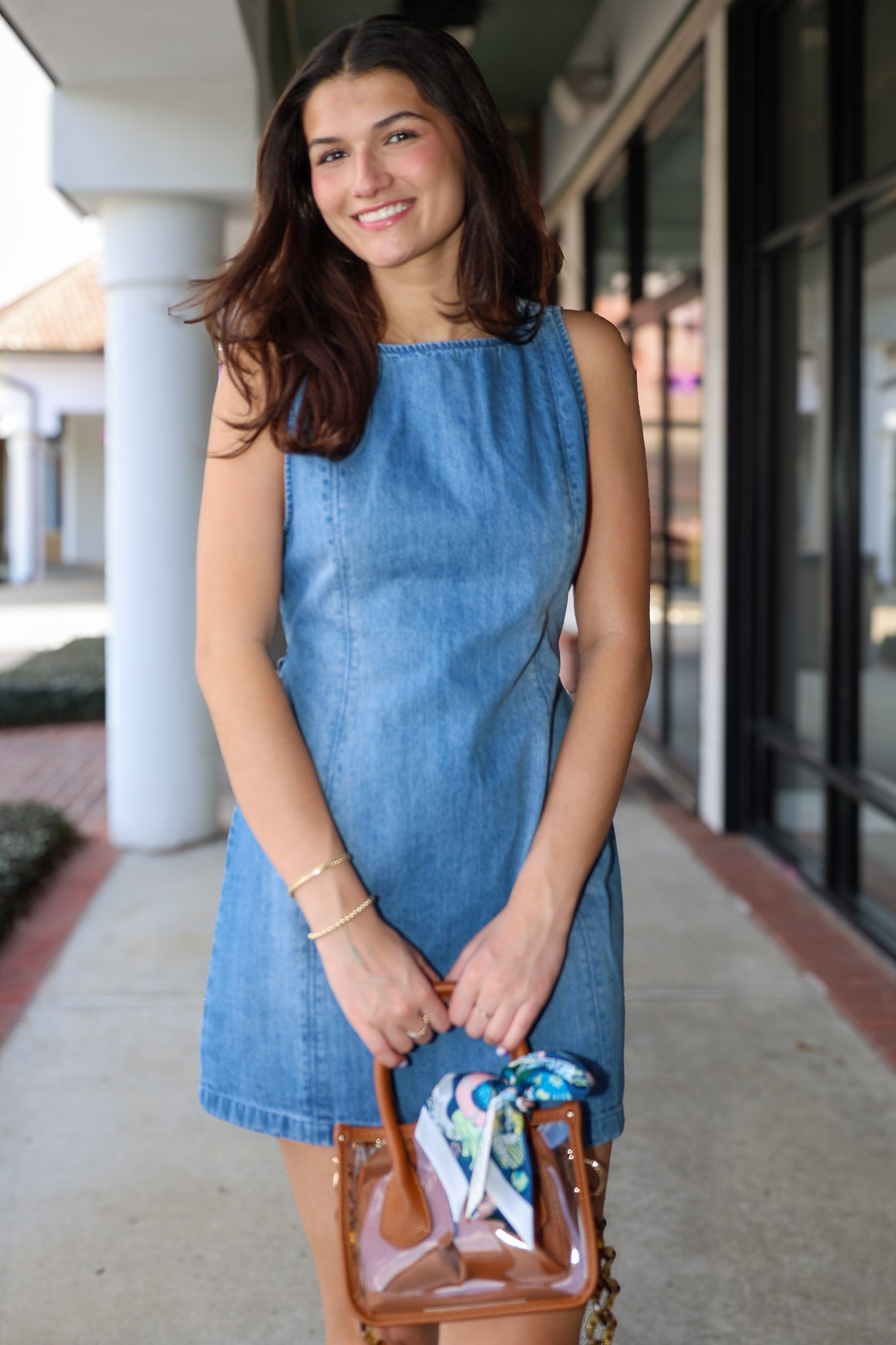 Woman wearing a blue denim dress holding a transparent handbag with a colorful interior.