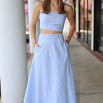 Woman wearing a light blue and white top and midi skirt standing on a sidewalk.
