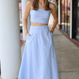 Woman wearing a light blue and white top and midi skirt standing on a sidewalk.