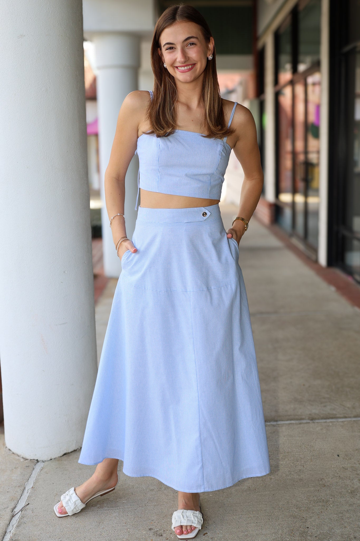 Woman wearing a light blue and white top and midi skirt standing on a sidewalk.