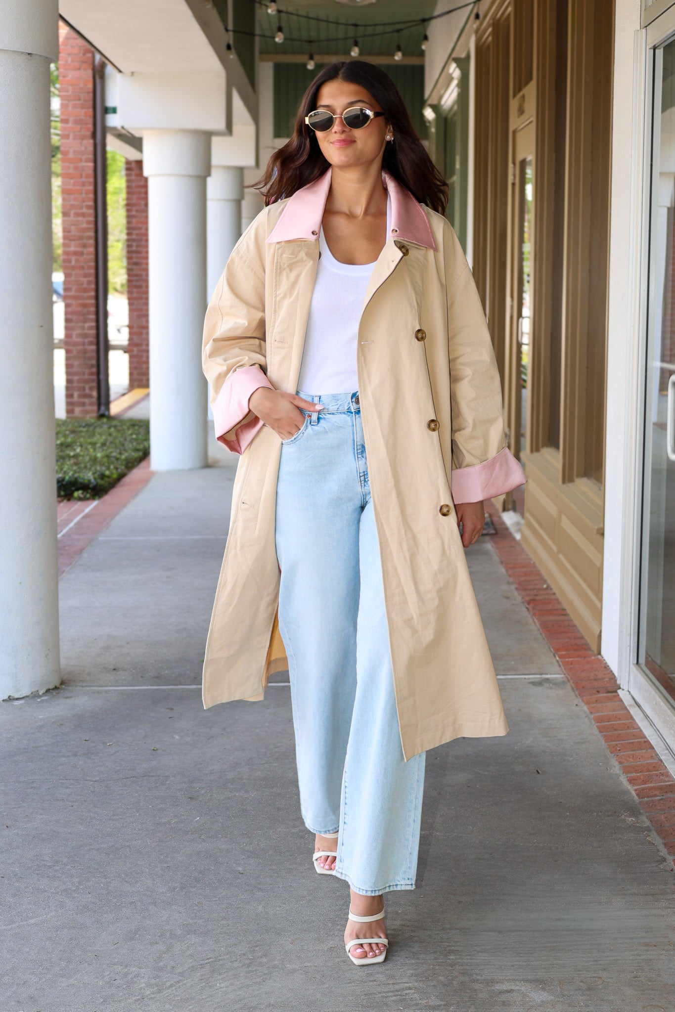 Woman wearing a beige trench coat with pink lining, white top, and light blue jeans on a sidewalk.