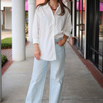 Bea Shirt, NIA - white button down shirt and light blue jeans standing on a sidewalk.