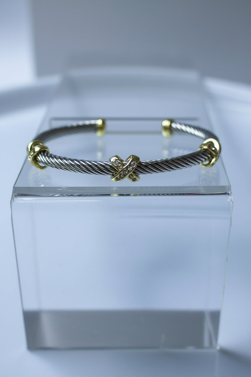 A silver-toned cable-wrapped cuff bracelet with gold-toned accents, displayed on a clear acrylic block against a light gray background.