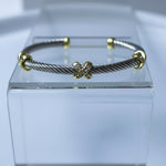 A silver-toned cable-wrapped cuff bracelet with gold-toned accents, displayed on a clear acrylic block against a light gray background.