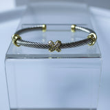 A silver-toned cable-wrapped cuff bracelet with gold-toned accents, displayed on a clear acrylic block against a light gray background.
