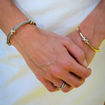 A close-up of a person's hands clasped in front of a white skirt, wearing a silver cable-style bracelet on the left wrist and a gold star-shaped bangle on the right wrist.