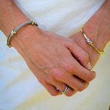 A close-up of a person's hands clasped in front of a white skirt, wearing a silver cable-style bracelet on the left wrist and a gold star-shaped bangle on the right wrist.
