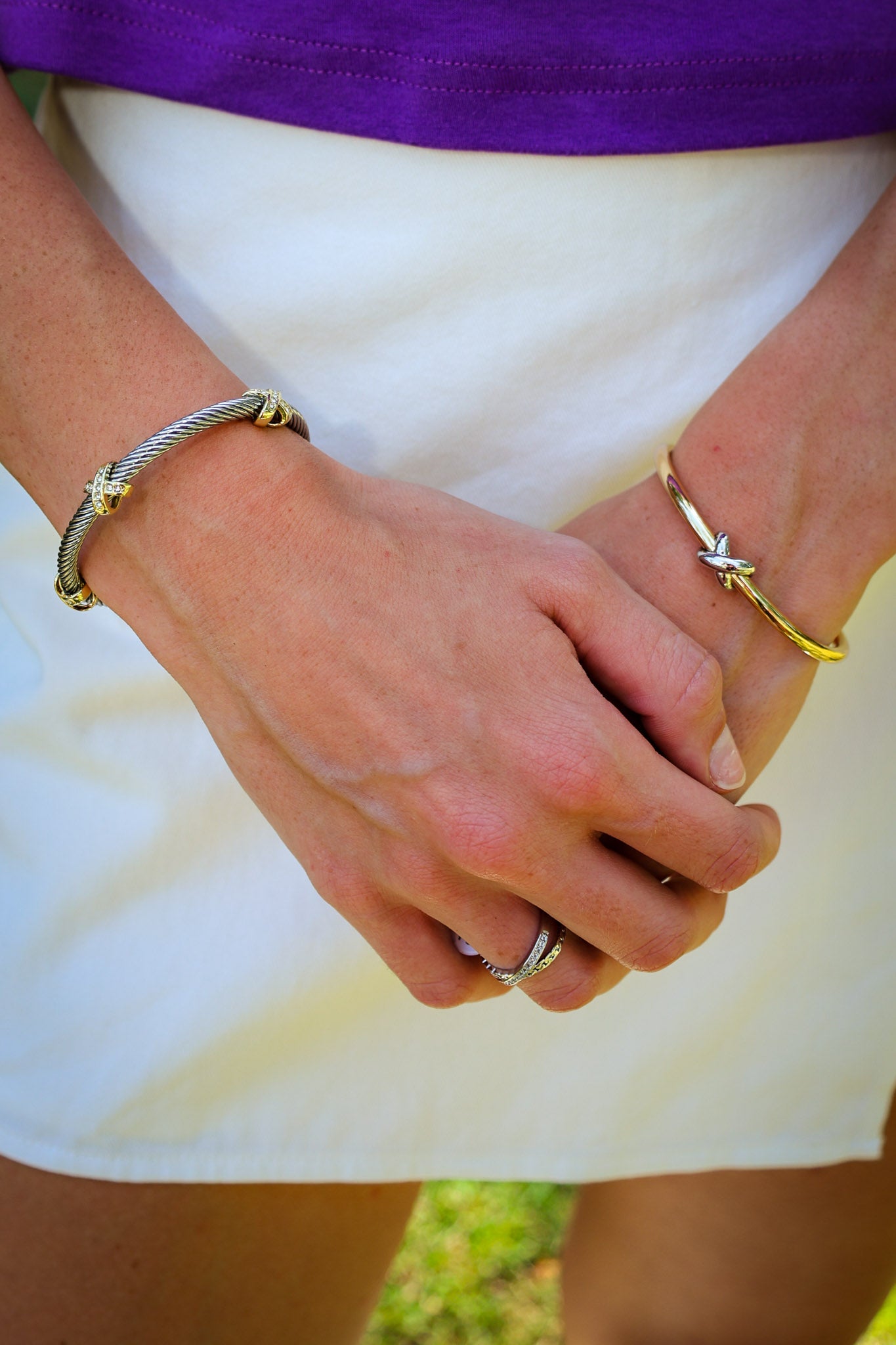 A close-up of a person's hands clasped in front of a white skirt, wearing a silver cable-style bracelet on the left wrist and a gold star-shaped bangle on the right wrist.