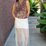 A woman stands outdoors on a sidewalk wearing a sleeveless brown sheer mesh top with white polka dots and a white semi-sheer midi skirt, paired with brown ankle boots.