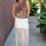 A woman stands outdoors on a sidewalk wearing a sleeveless brown sheer mesh top with white polka dots and a white semi-sheer midi skirt, paired with brown ankle boots.