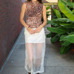 A woman stands on a sidewalk wearing a sheer brown mesh top with white polka dots, a white semi-sheer midi skirt, and brown leather ankle boots. She is posed between a red brick wall and large green tropical plants.