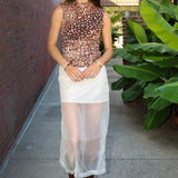 A woman stands on a sidewalk wearing a sheer brown mesh top with white polka dots, a white semi-sheer midi skirt, and brown leather ankle boots. She is posed between a red brick wall and large green tropical plants.