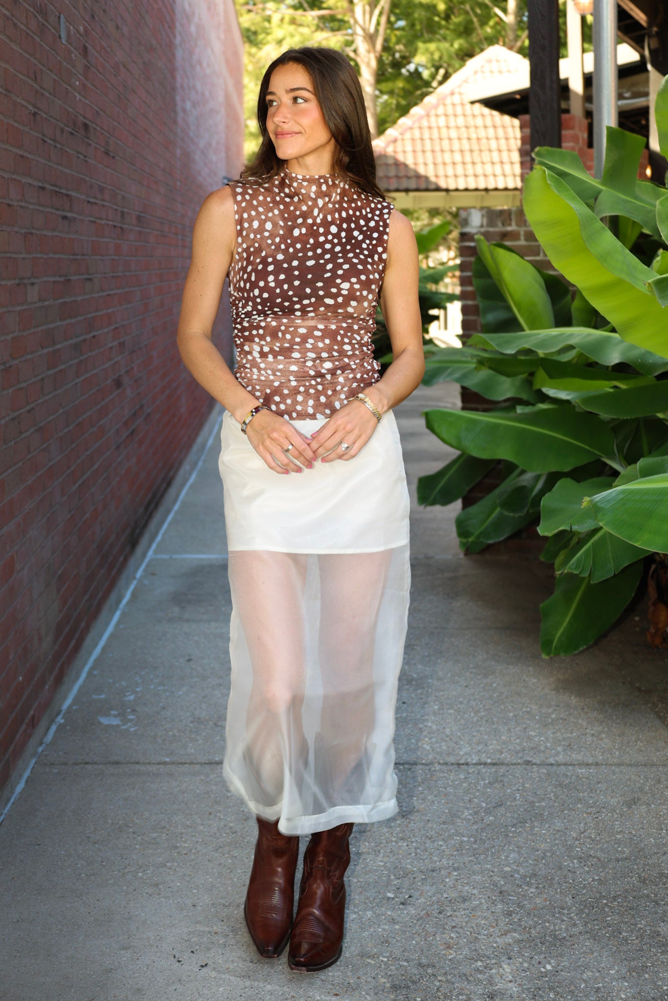 A woman stands on a sidewalk wearing a sheer brown mesh top with white polka dots, a white semi-sheer midi skirt, and brown leather ankle boots. She is posed between a red brick wall and large green tropical plants.