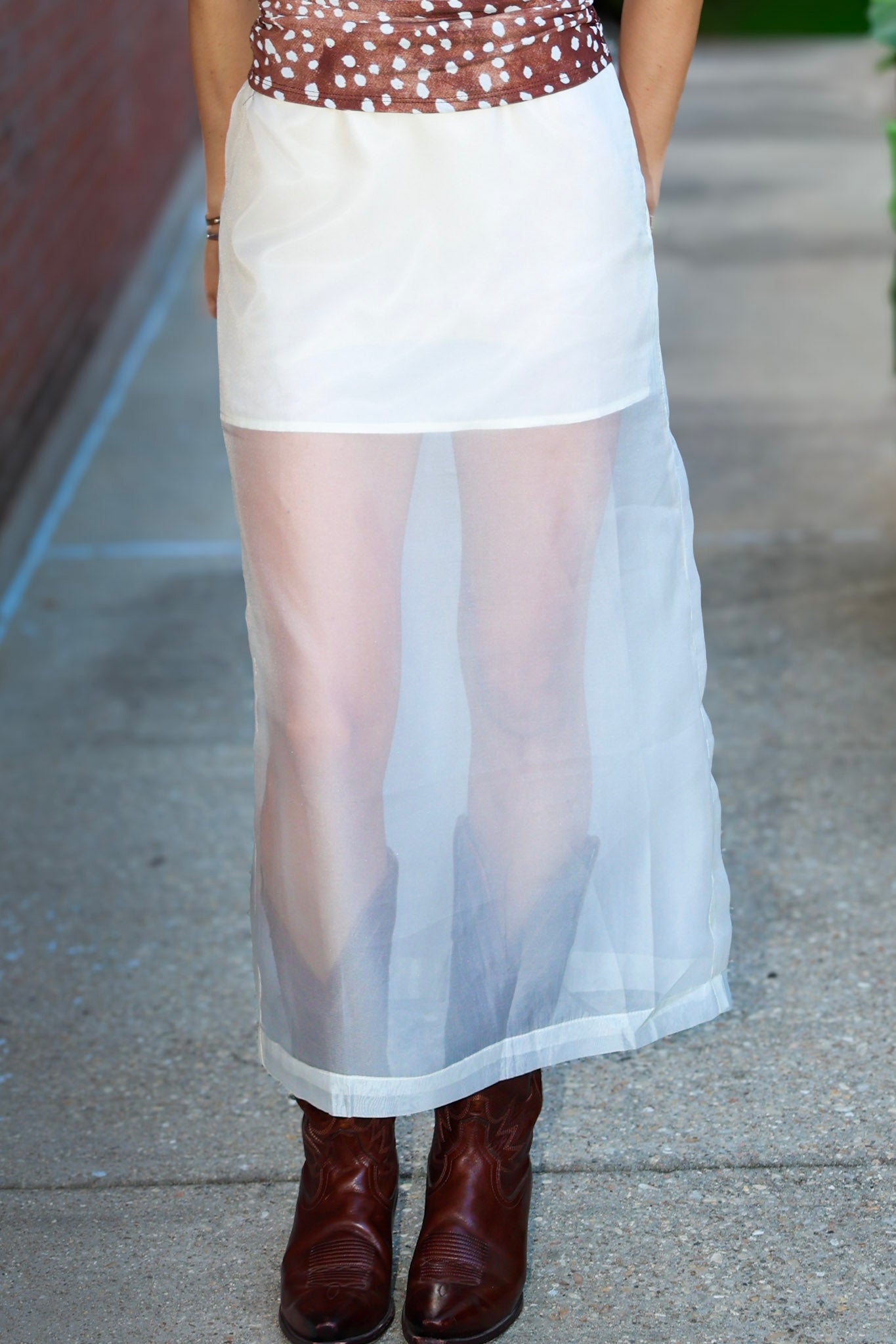 A person wearing a white semi-sheer midi skirt with a brown polka-dot top and brown leather cowboy boots, standing on a concrete sidewalk.