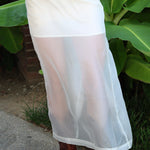 A white sheer mesh maxi skirt worn by a model, paired with brown leather cowboy boots, standing outdoors in front of green banana leaves.