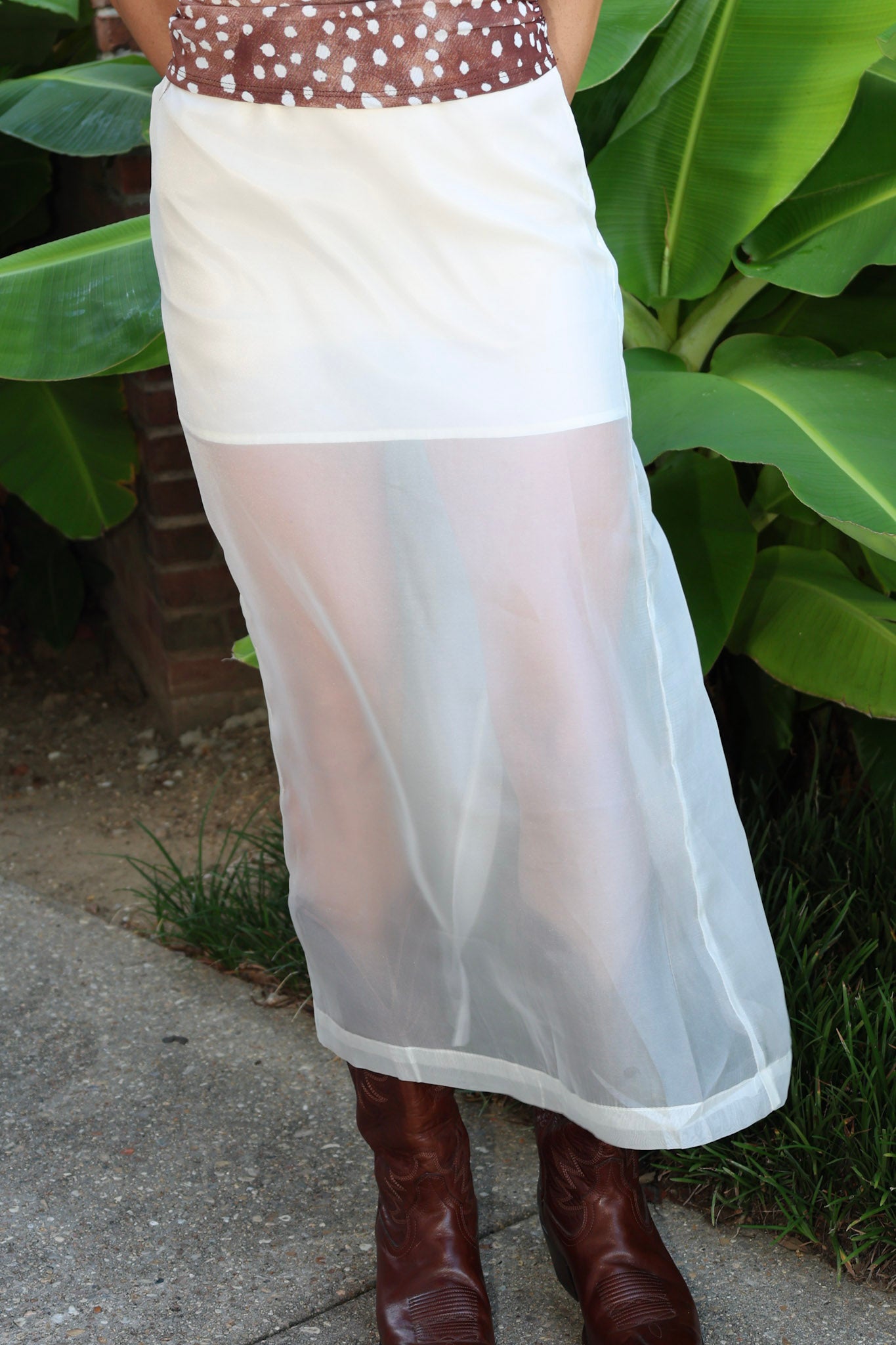 A white sheer mesh maxi skirt worn by a model, paired with brown leather cowboy boots, standing outdoors in front of green banana leaves.